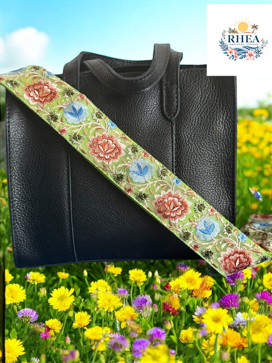 Close-up of a handmade crossbody strap in seafoam green with embroidered red, blue, and green floral motifs. Styled on a black leather tote against a meadow backdrop. Part of the Meadow Bloom Collection by Rhea Artistry Studio.