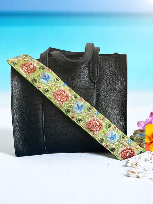 Close-up of a handmade crossbody strap in seafoam green with embroidered red, blue, and green floral motifs. Styled on a black leather tote against a tropical beach backdrop. Part of the Meadow Bloom Collection by Rhea Artistry Studio.
