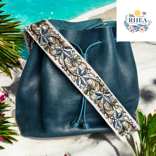 2-inch wide embroidered strap with blue and olive florals on an ivory base, styled on a deep teal leather bucket bag on a beach setting.