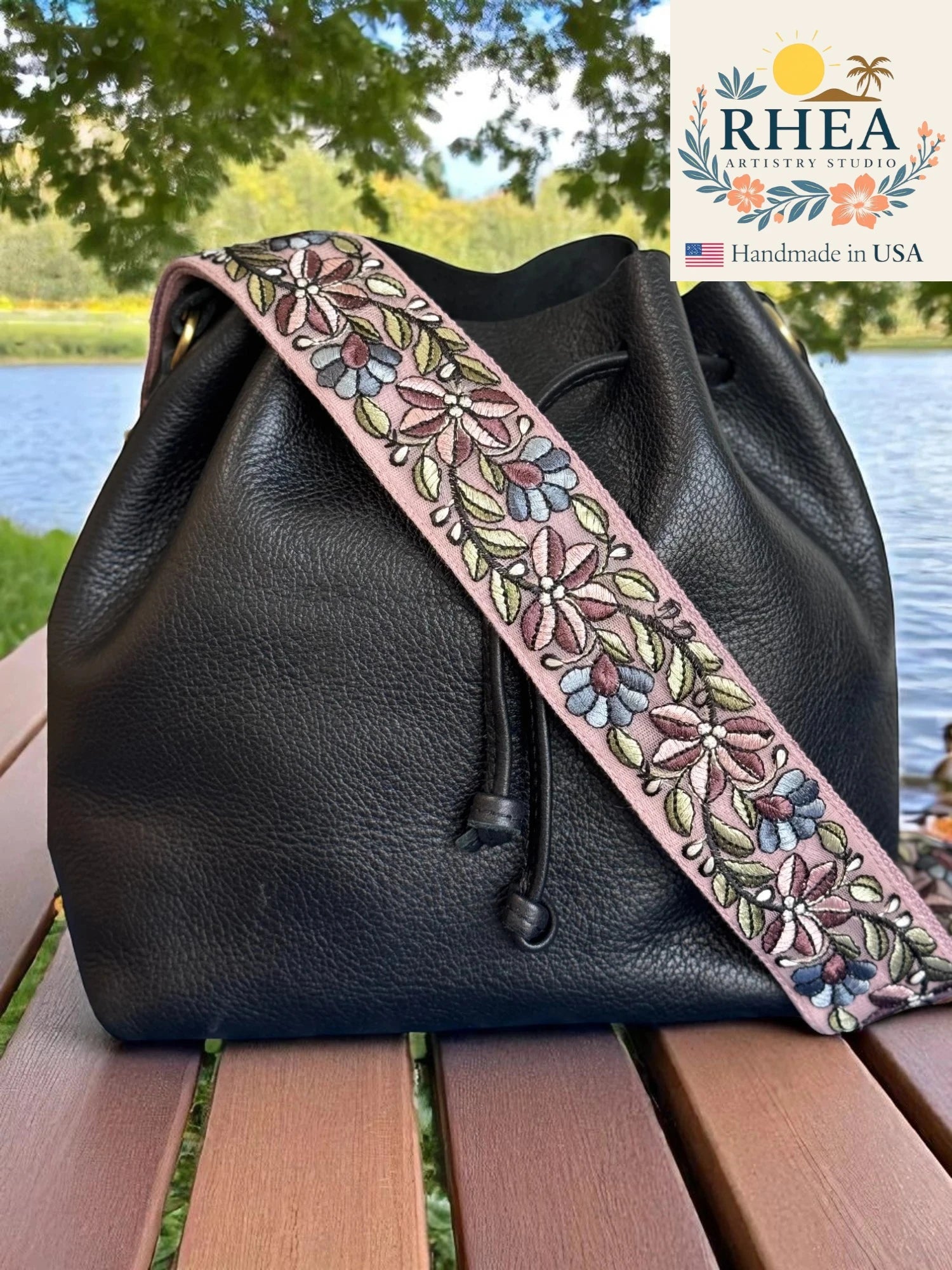Black leather bag with a floral purse strap on a wooden surface by a lake, with RHEA branding.