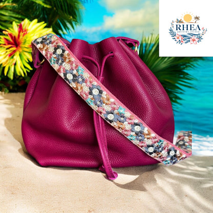 A richly embroidered strap featuring teal, navy, bronze, pink, and white floral motifs on cream webbing, styled with a magenta leather bag at the beach