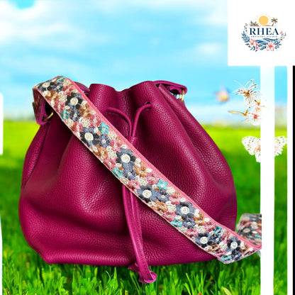  A richly embroidered strap featuring teal, navy, bronze, pink, and white floral motifs on cream webbing, styled with a magenta leather bag in the garden setting with butterflies around it