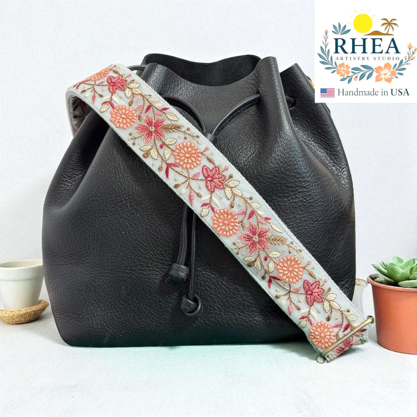 Black leather bag with a floral embroidered strap on a light background, featuring the RHEA brand.