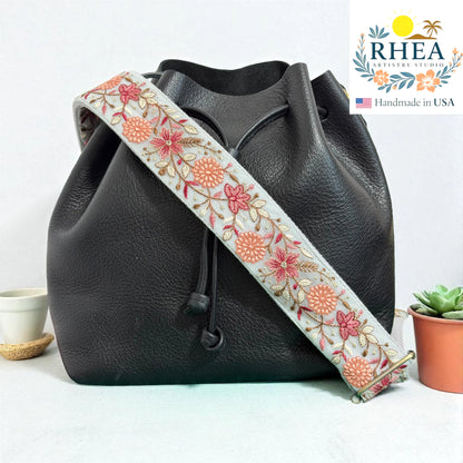 Black leather bag with a floral embroidered strap on a light background, featuring the RHEA brand.