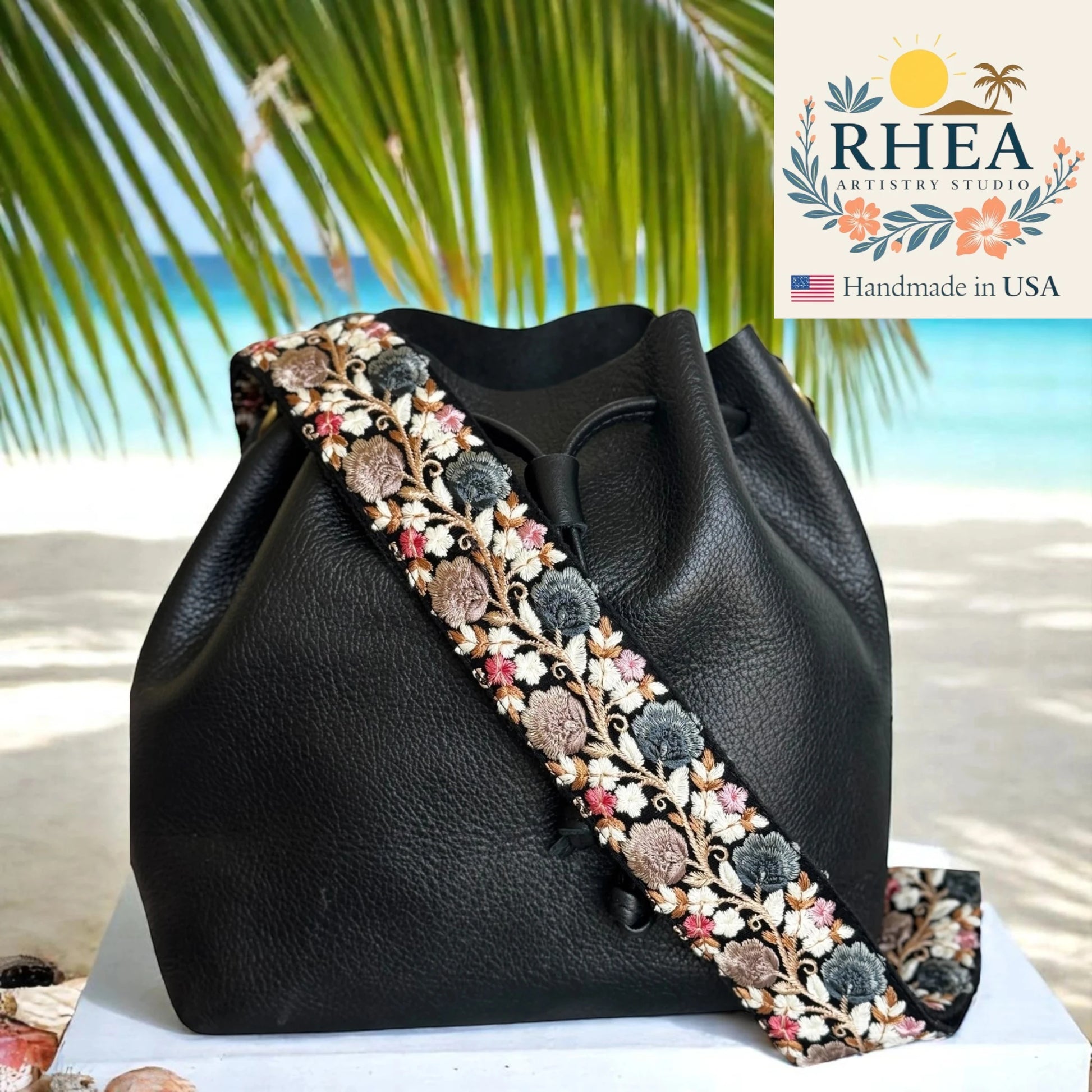 Black handbag with floral purse strap on a beach background, featuring RHEA Artisan Studio logo.
