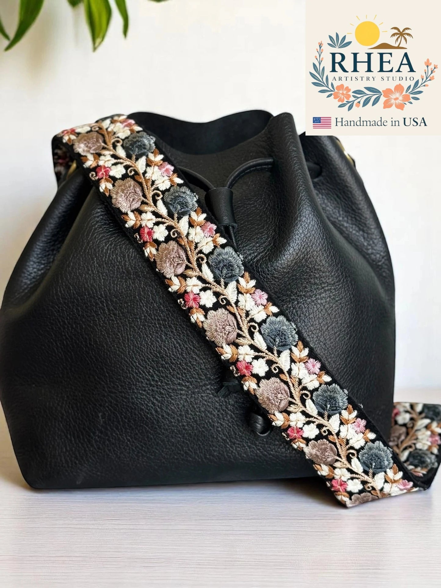 Black leather bag with floral embroidery bag strap on a white background, featuring the RHEA brand.