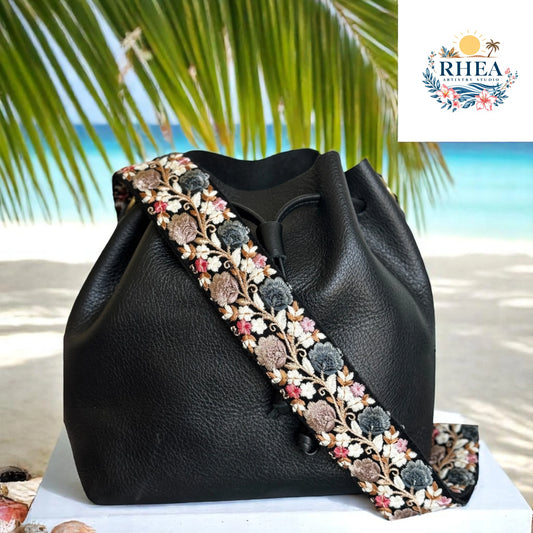 2-inch wide artisan strap with dusty pink, blue, and beige florals embroidered on black, styled on a black leather bag against a beach setting