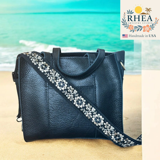 Black leather handbag with floral strap on a beach, featuring the RHEA brand.