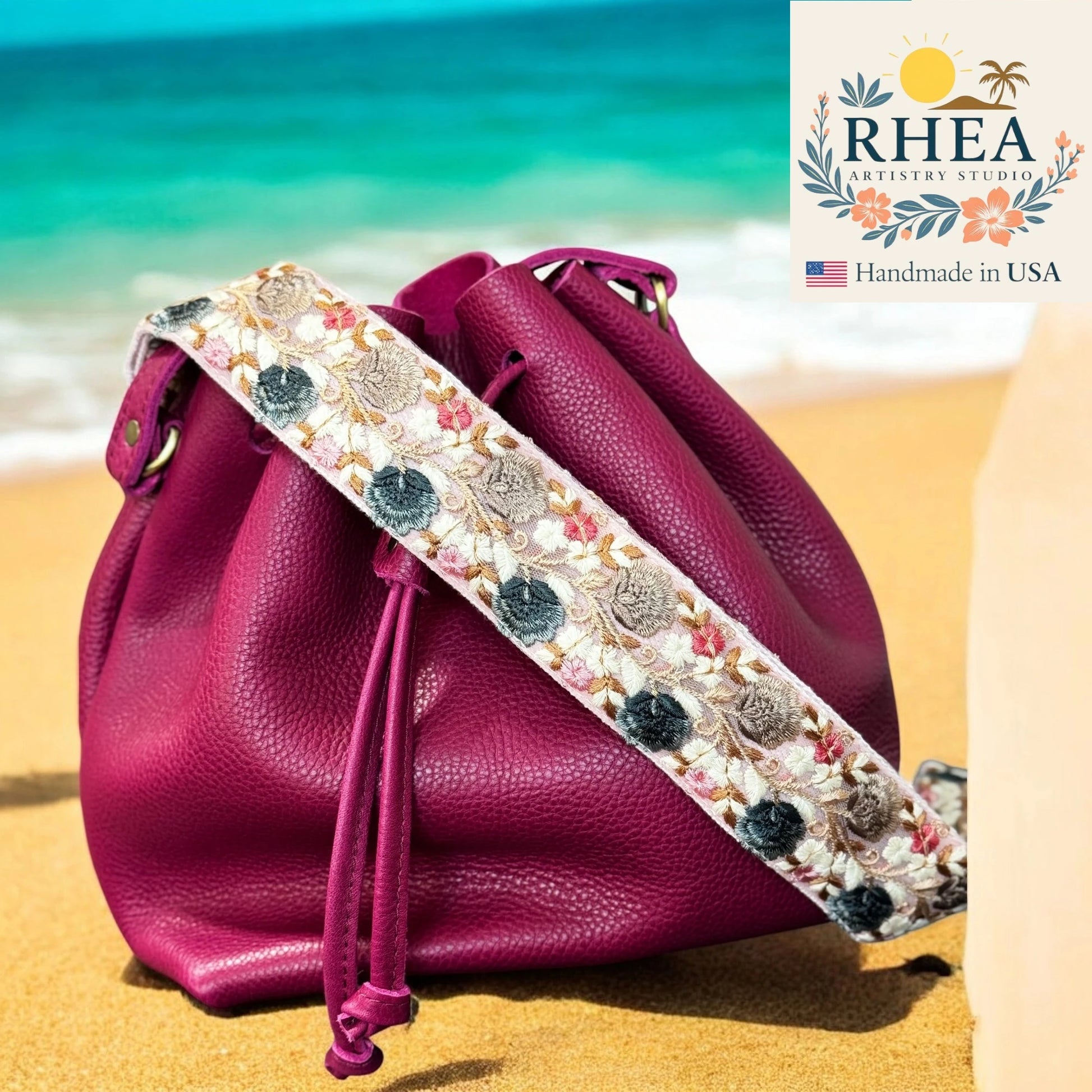 Pink leather bag with an embroidered purse strap on a beach, featuring the RHEA Artistry Studio logo.