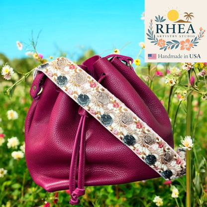 Purple drawstring bag with floral purse strap in a field of flowers, featuring RHEA Artistry Studio logo.
