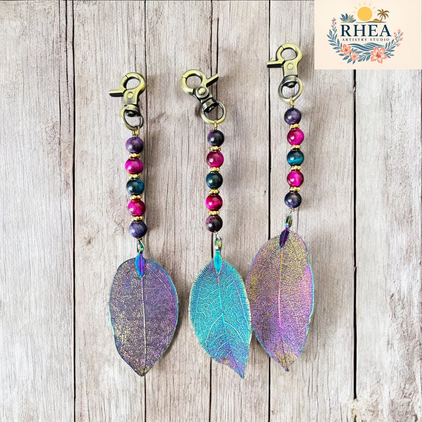 Three colorful leaf-shaped keychains on a wooden surface with 'RHEA' branding.