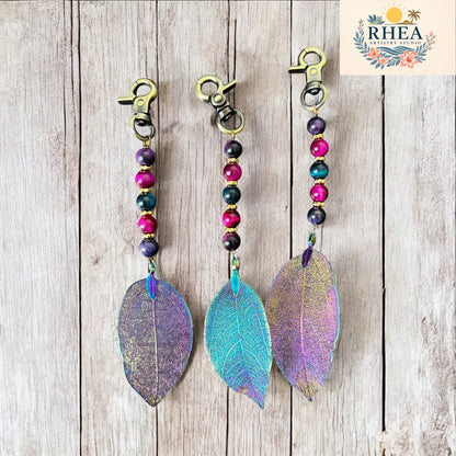 Three colorful leaf-shaped keychains on a wooden surface with 'RHEA' branding.