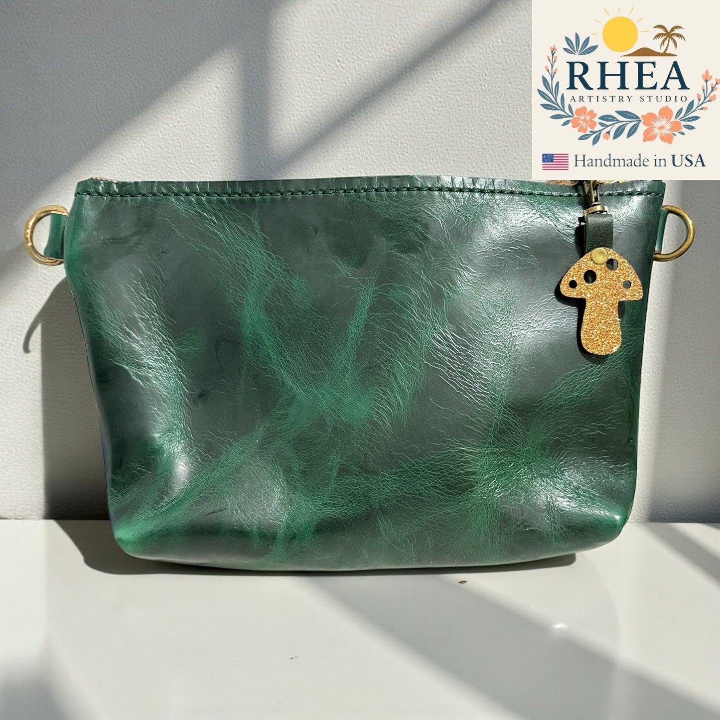 Green leather pouch with a gold mushroom charm on a white surface, featuring the RHEA Antiquary Studios logo.