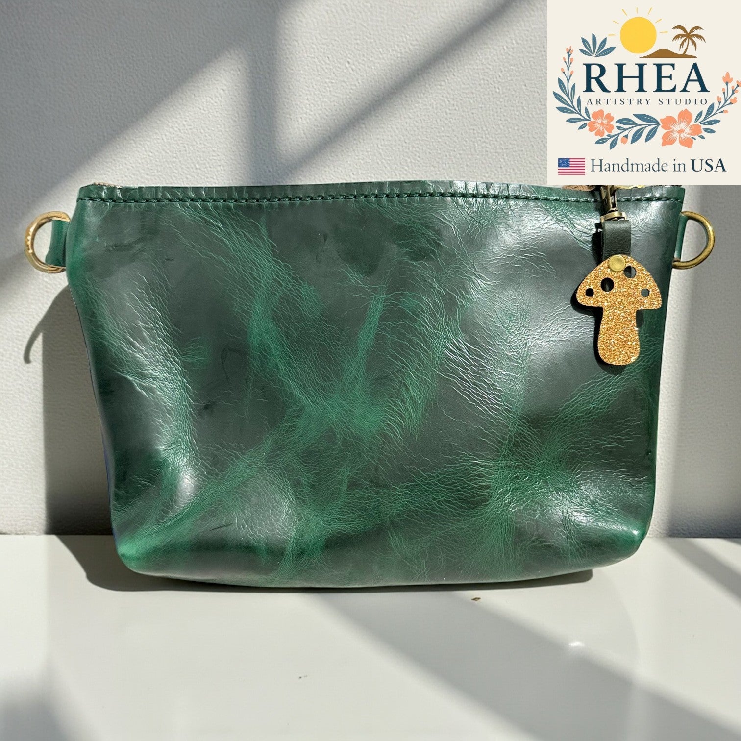 Green leather pouch with a gold mushroom charm on a white surface, featuring the RHEA Antiquary Studios logo.