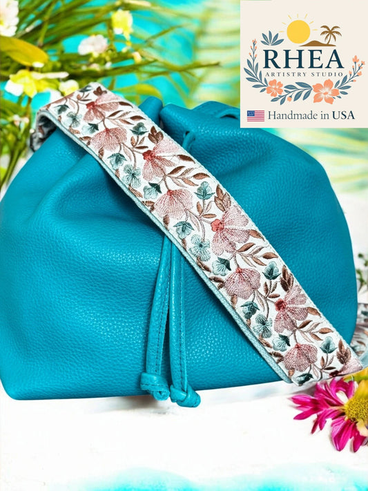 Blue handbag with a floral strap on a white background, featuring the RHEA Artistry Studio logo.