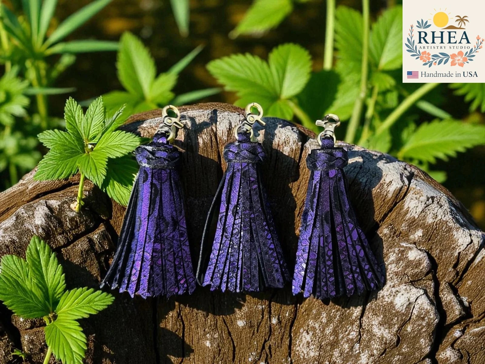 Three purple tassel earrings on a wooden block with green leaves in the background, featuring the RHEA brand.