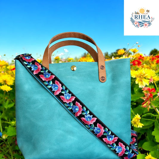 A handmade black adjustable strap with vivid floral embroidery in fuchsia pink, deep blue, and green, attached to a turquoise leather tote bag by the garden. From the Ocean Bloom Collection.