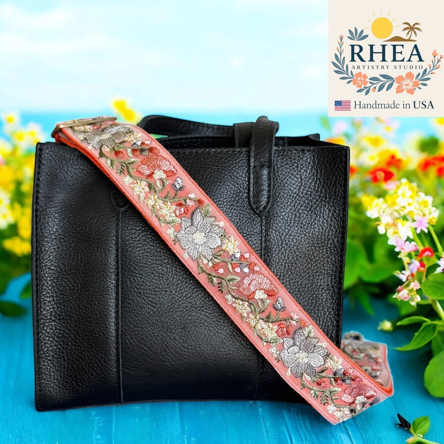 Black handbag with a floral embroidered purse strap on a blue surface with flowers in the background, featuring the RHEA Artistry Studio logo.