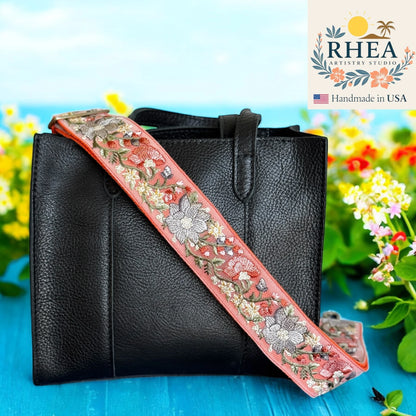 Black handbag with a floral embroidered purse strap on a blue surface with flowers in the background, featuring the RHEA Artistry Studio logo.