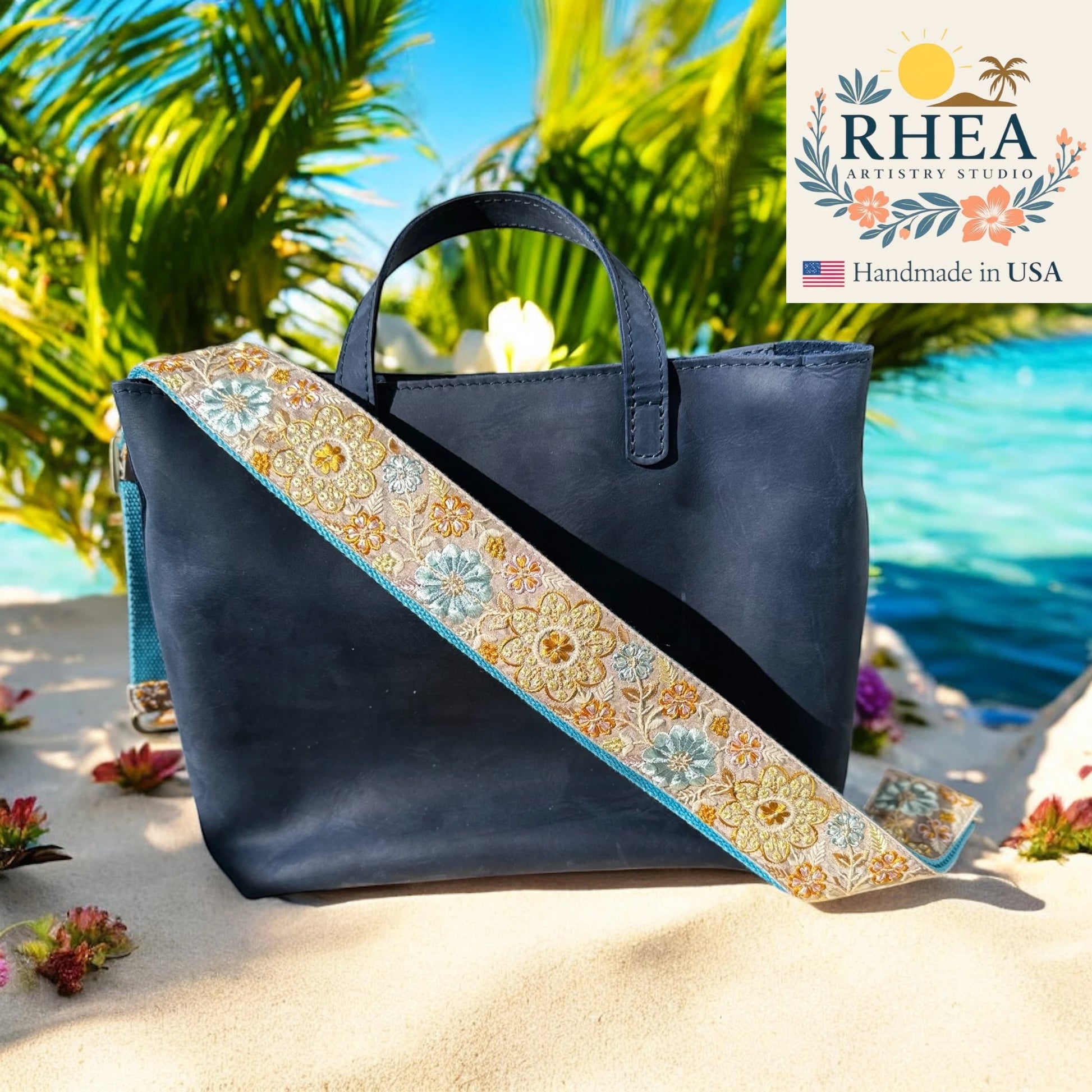 Navy blue handbag with a floral purse strap on a sandy beach with palm trees and water in the background, featuring the RHEA brand.