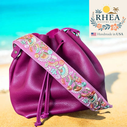 Pink drawstring bag with floral strap on a beach, featuring RHEA Artistry Studio logo.