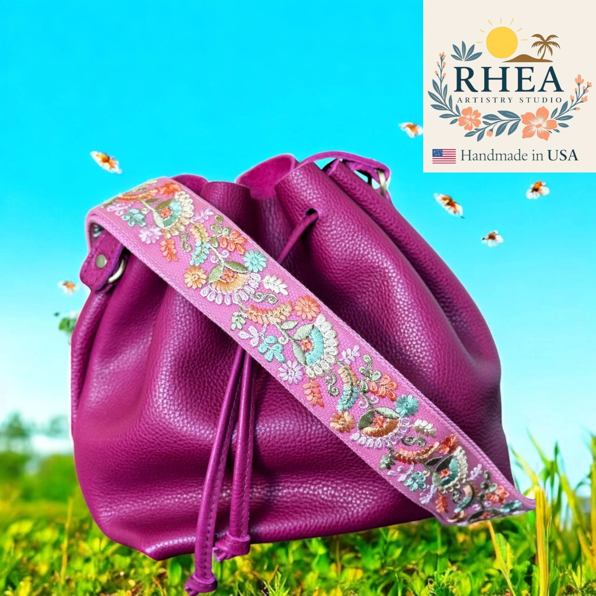 Purple drawstring bag with floral strap on grass with blue sky, RHEA brand visible