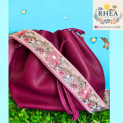 Pink leather bag with floral embroidery purse strap on a grassy background, featuring the RHEA brand.