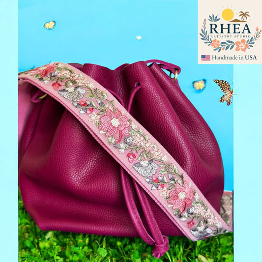Pink leather bag with floral embroidery purse strap on a grassy background, featuring the RHEA brand.