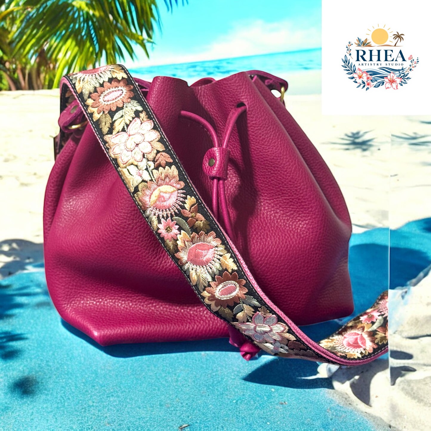 Fuchsia leather bucket bag with a richly embroidered crossbody strap featuring floral patterns in blush pink, ivory, coral, and golden olive, resting on a tropical beach backdrop with palm trees and ocean waves.