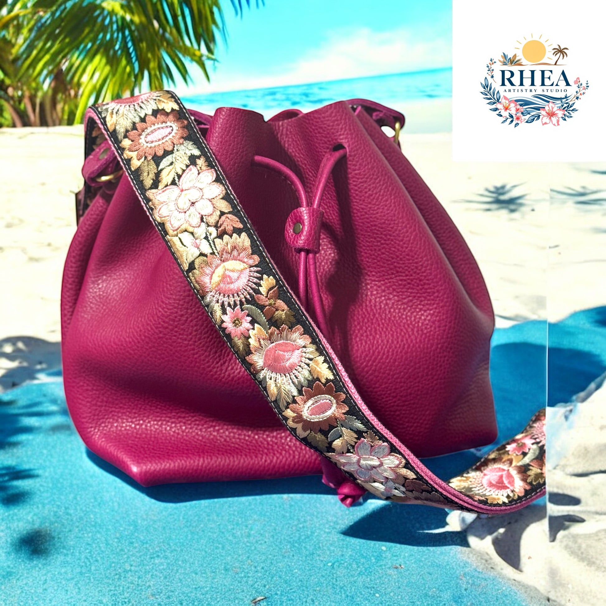 Fuchsia leather bucket bag with a richly embroidered crossbody strap featuring floral patterns in blush pink, ivory, coral, and golden olive, resting on a tropical beach backdrop with palm trees and ocean waves.