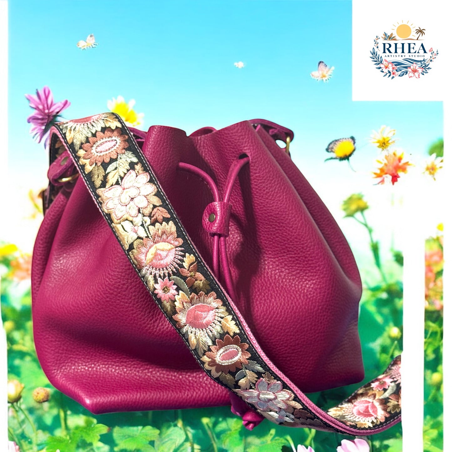 Fuchsia leather bucket bag with a richly embroidered crossbody strap featuring floral patterns in blush pink, ivory, coral, and golden olive, resting on a meadow garden