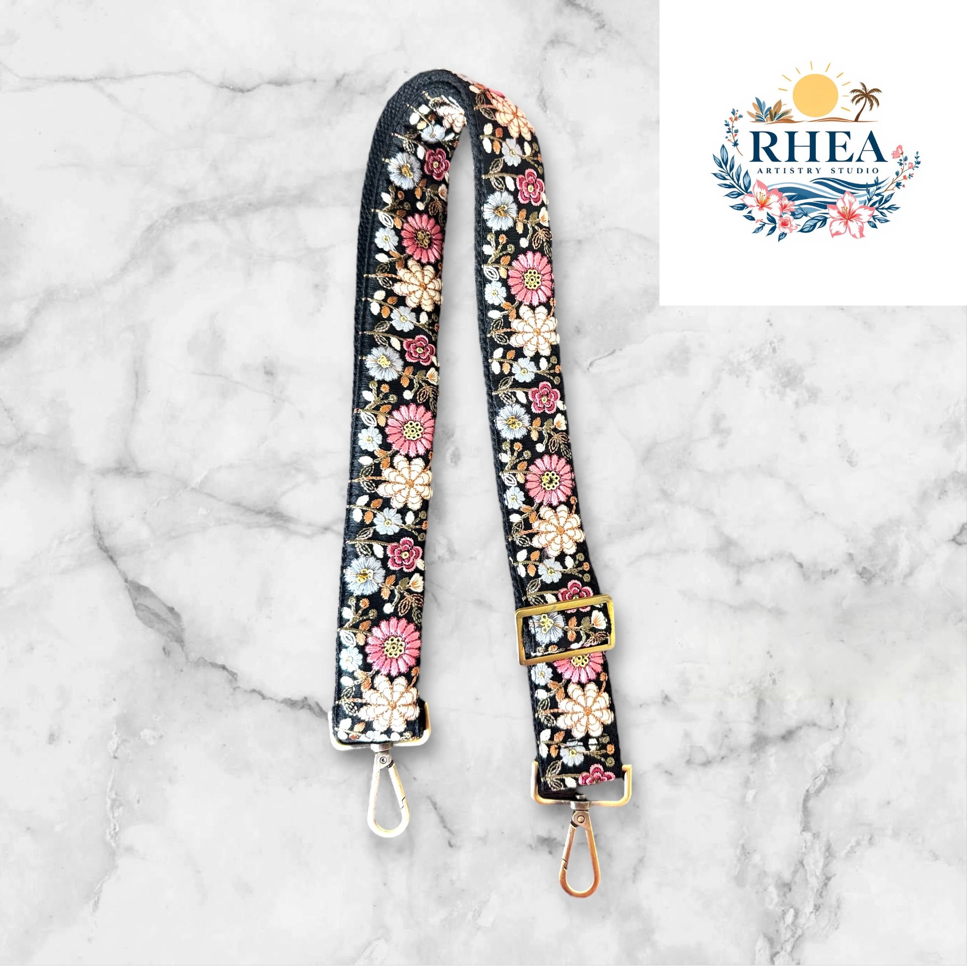 An adjustable strap with daisy flowers embroidery attached to a black bag, with a field and sunset in the background.