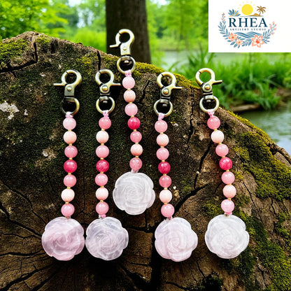 Pink beaded keychains with floral charms on a wooden log outdoors, featuring the Rhea brand logo.