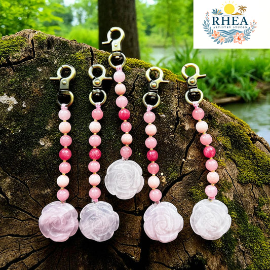 Pink beaded keychains with floral charms on a wooden log outdoors, featuring the Rhea brand logo.