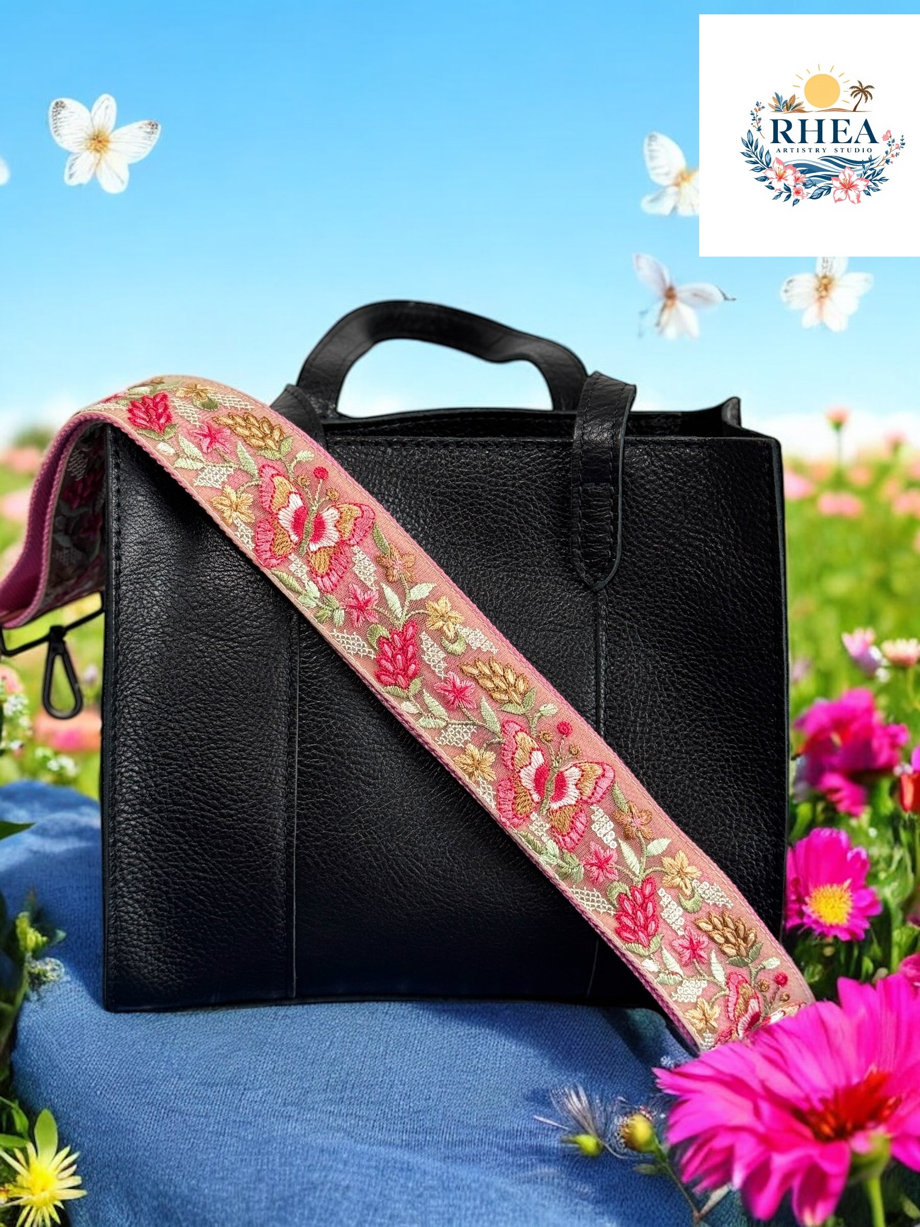 A black purse with a pink and green floral embroidered strap placed on a blue surface with flowers in the background.