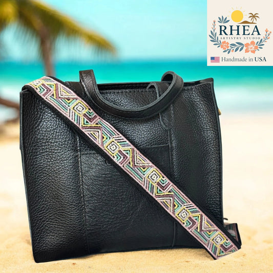 Black leather handbag with a colorful strap on a beach background, featuring the RHEA brand.