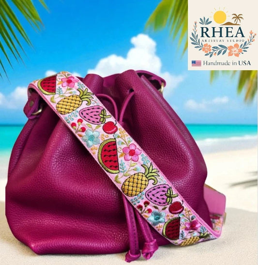 Pink leather bag with colorful fruit strap on a beach background, featuring the Rhea Artistry Studio logo.