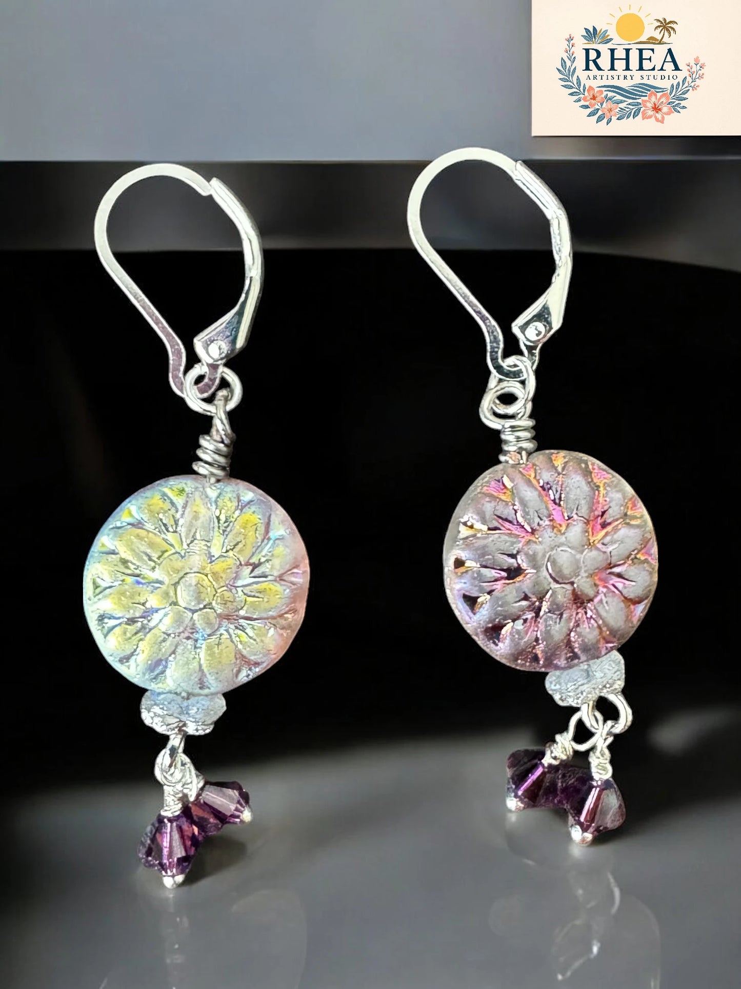 Pair of floral earrings with purple accents on a reflective surface, branded 'RHEA'.