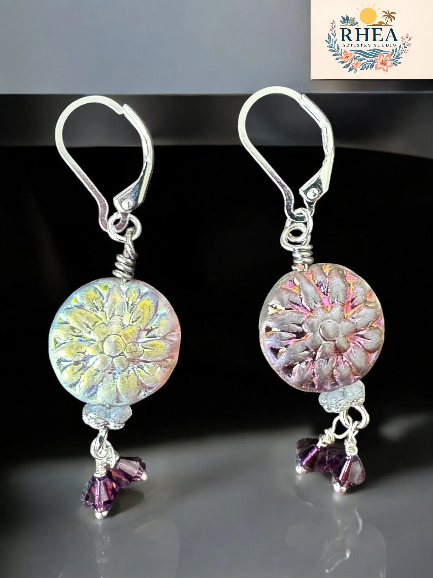 Pair of floral earrings with purple accents on a reflective surface, branded 'RHEA'.