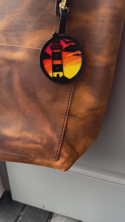 A hand-painted leather charm with a lighthouse design, attached to a brown leather bag with a brass clasp.
