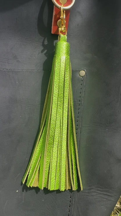 A jumbo leather tassel accessory in fern green color, displayed on a black handbag with a red strap.