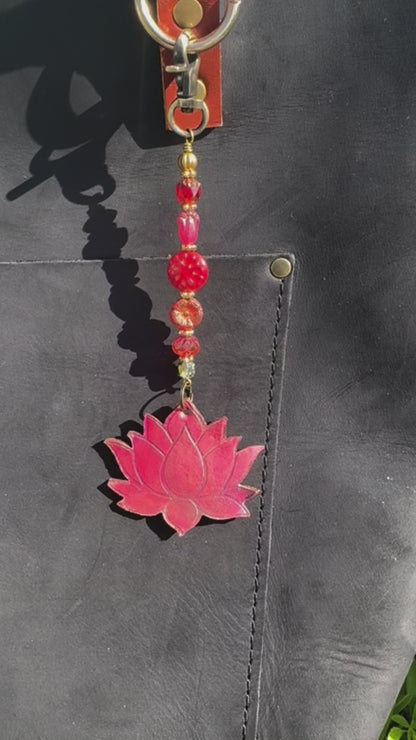 A handcrafted lotus bloom charm made of red and orange glass with brass hardware, attached to a leather-bound notebook.