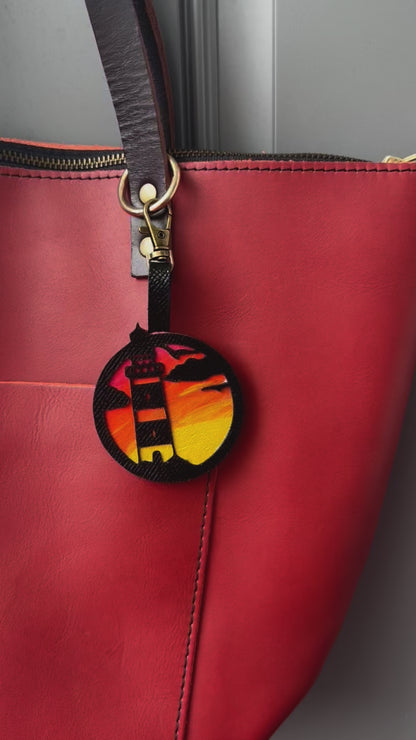 A hand-painted leather charm with a lighthouse design, attached to a red leather bag with a brass clasp.