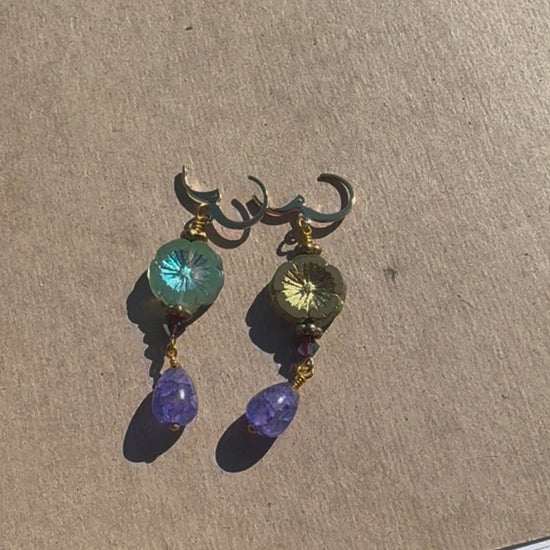A pair of hibiscus violet drop earrings with gemstones and crystal embellishments, placed on a brown surface.