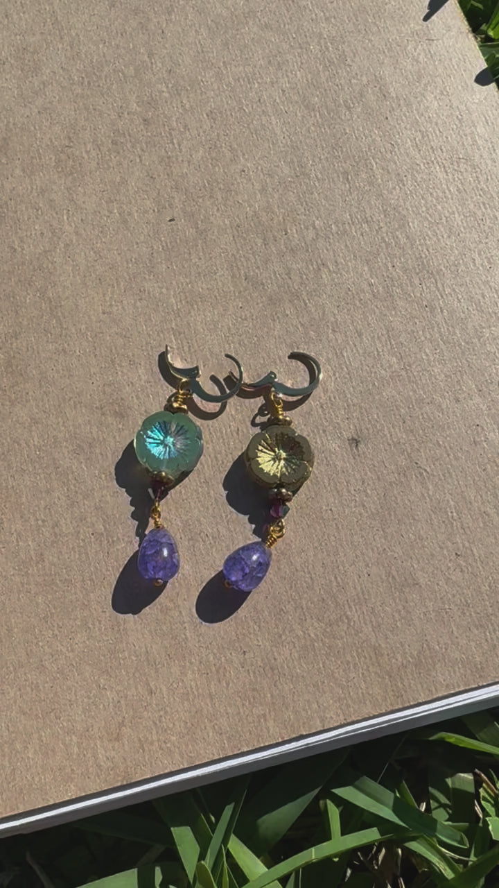 A pair of hibiscus violet drop earrings with gemstones and crystal embellishments, placed on a brown surface.