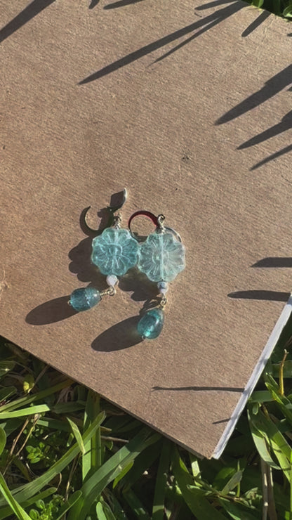 A pair of Daisy Blue Silver Drop Earrings with a blue gemstone, displayed on a brown cardboard surface outdoors.