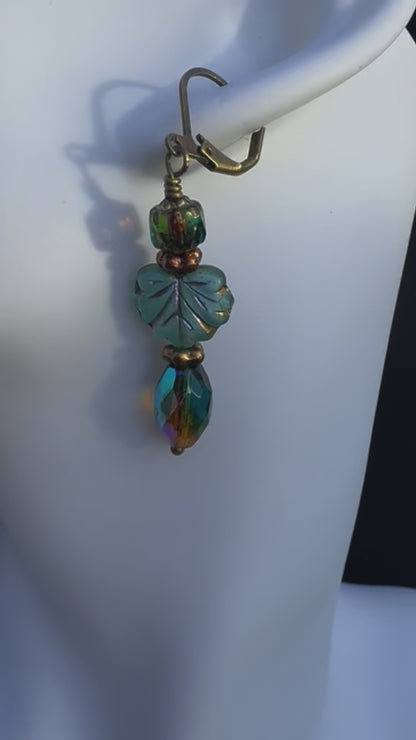 A pair of handcrafted earrings with green and blue czech glass beads, antique bronze finish, and brass hardware on a textured background.