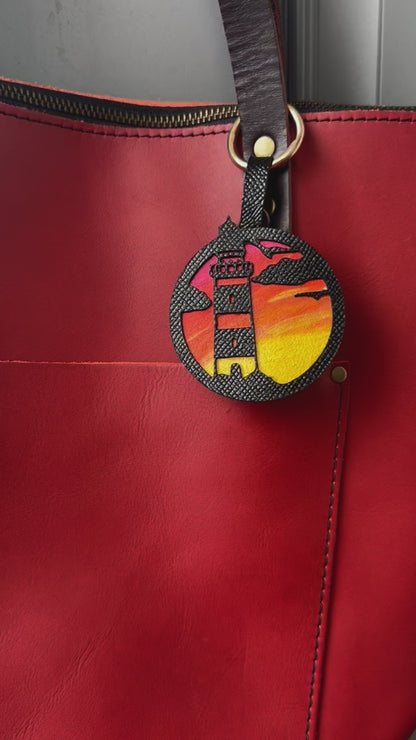 A hand-painted leather charm with a lighthouse design, attached to a red leather bag with a brass clasp.