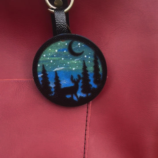 A handpainted leather charm with a starry night landscape, a deer, and a forest design, attached to a red leather bag.