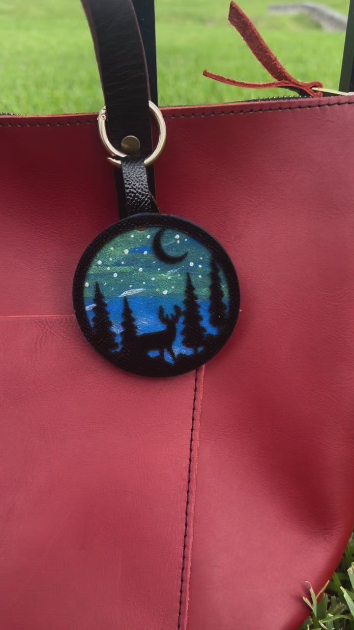 A handpainted leather charm with a starry night landscape, a deer, and a forest design, attached to a red leather bag.